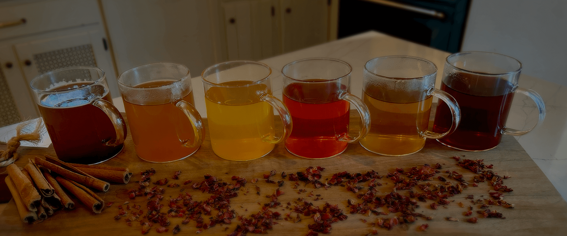 Six glasses of the different Sync Botanicals teas lined up on a table with loose herbs.
