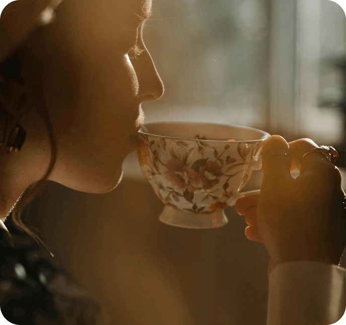 Person sipping tea from a floral teacup in warm window light.